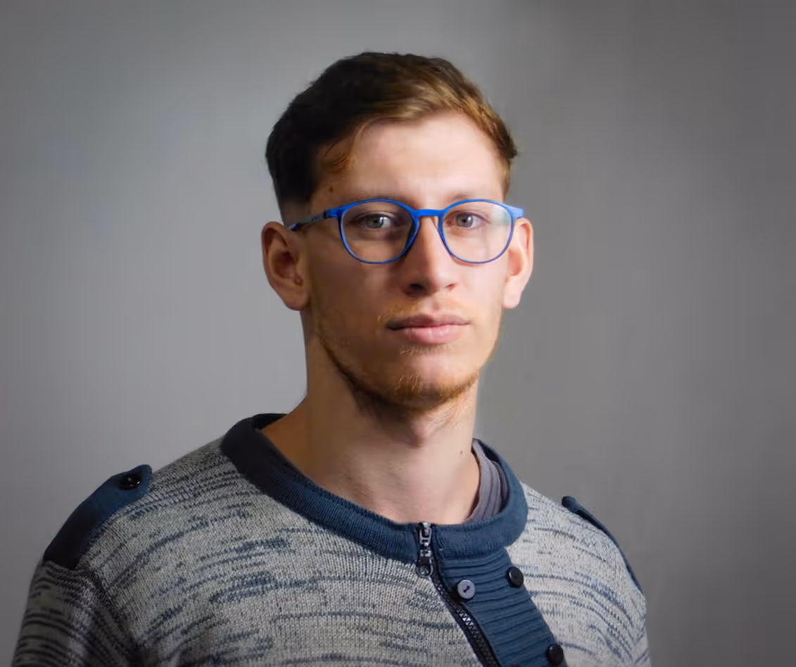 Young man with short brown hair and blue glasses wearing a gray and blue knit sweater looking at the camera.